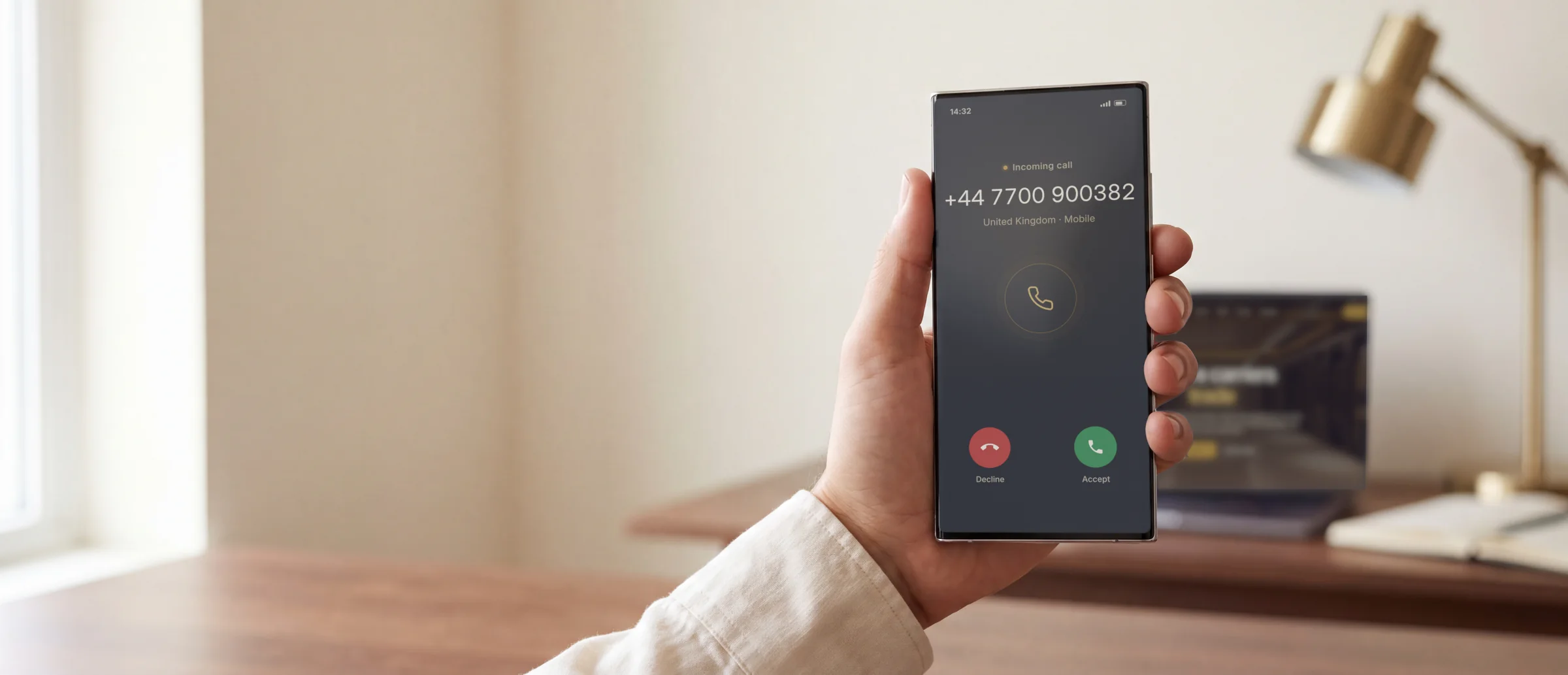 A hand holding a phone showing an incoming call from a UK number, a laptop on the desk behind
