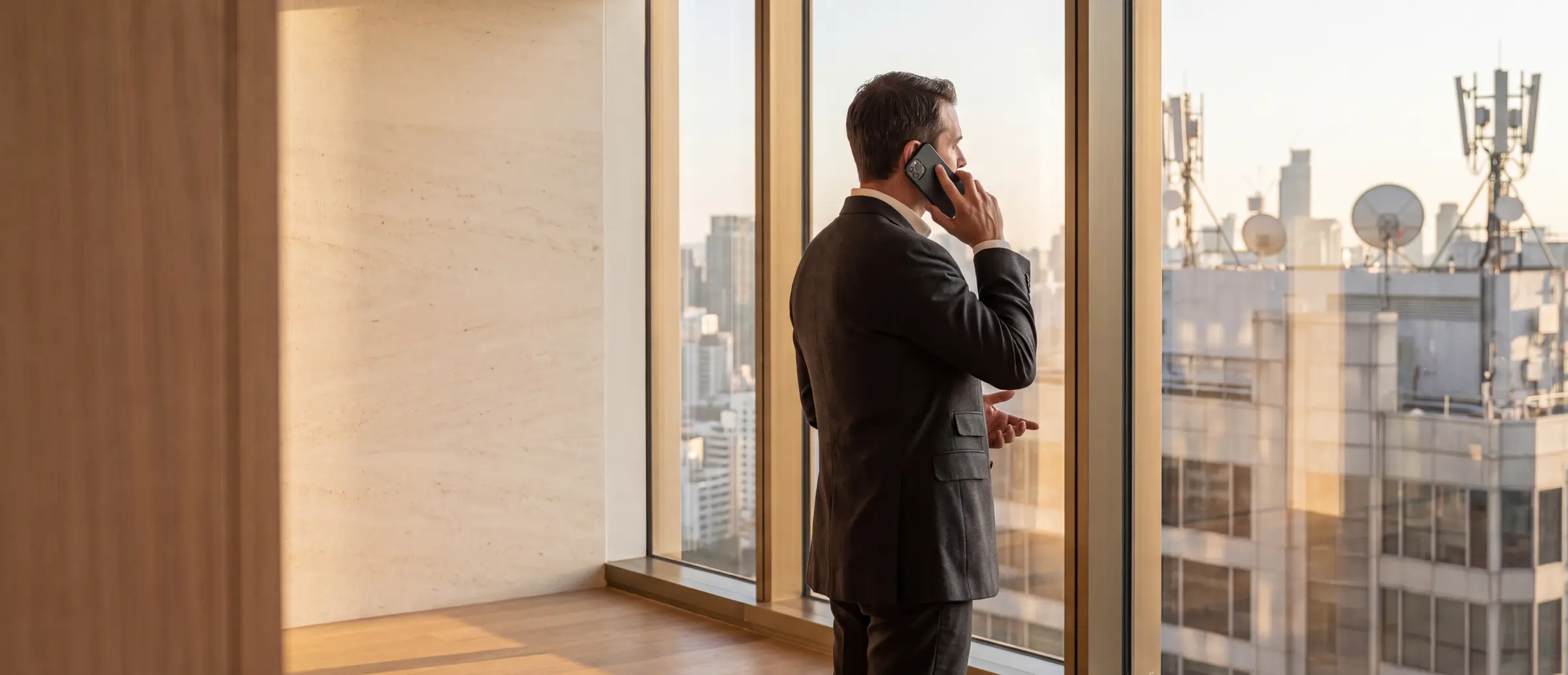 A businessman on a phone call at a high-floor window, rooftop telecom antennas across the skyline